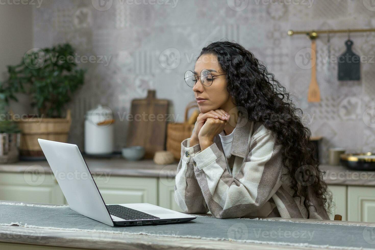 Hispano mujer pensando a hogar sentado en cocina trabajando en computadora portátil, persona de libre dedicación en lentes y Rizado cabello, mujer de negocios trabajando y aprendizaje en línea remotamente foto
