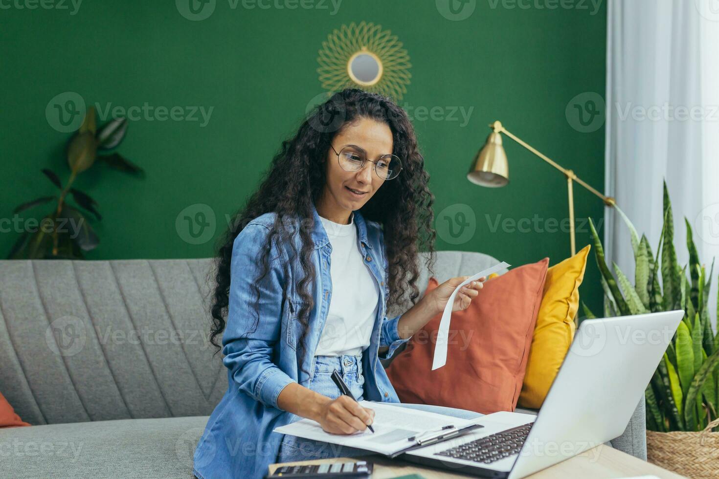 Young beautiful hispanic woman student studying online at home from laptop. Sits on the sofa, writes, listens to a lecture, passes an exam. photo