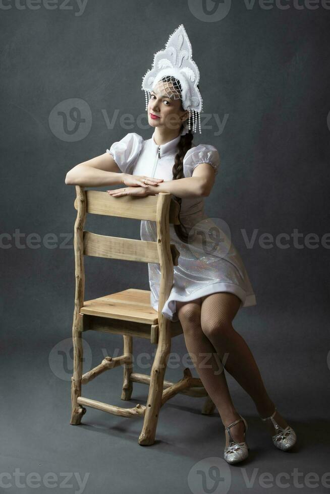 Portrait of a beautiful girl in a snow maiden costume on a gray background. A Russian snow girl in a crown sits on a wooden chair. photo