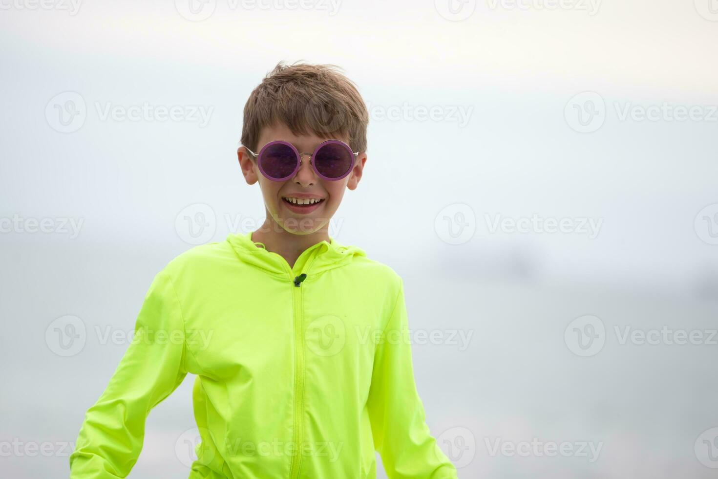 A boy in a bright green sweater and purple glasses looks at the camera, smiles. photo