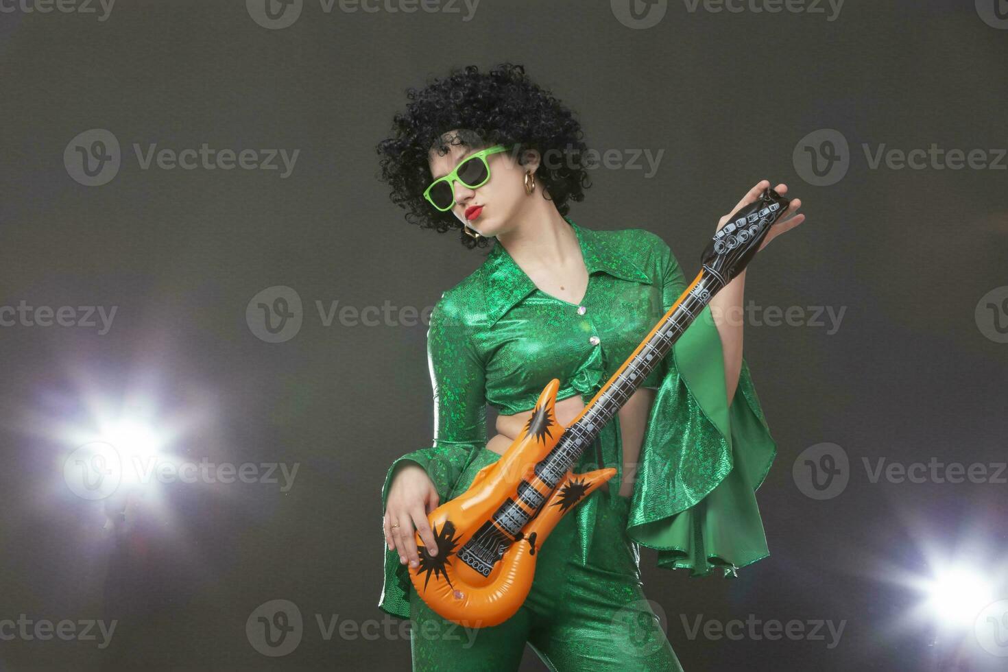 A disco-style girl in a green suit and a toy guitar posing on a gray background. photo