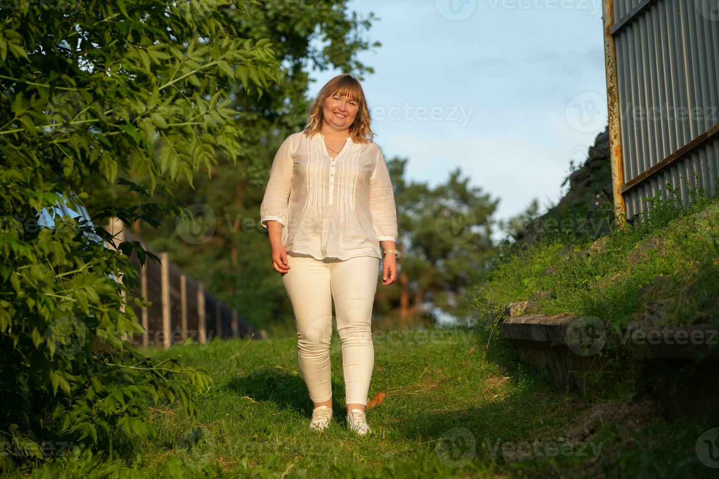 A beautiful blond woman with a full build walks in a green park. photo