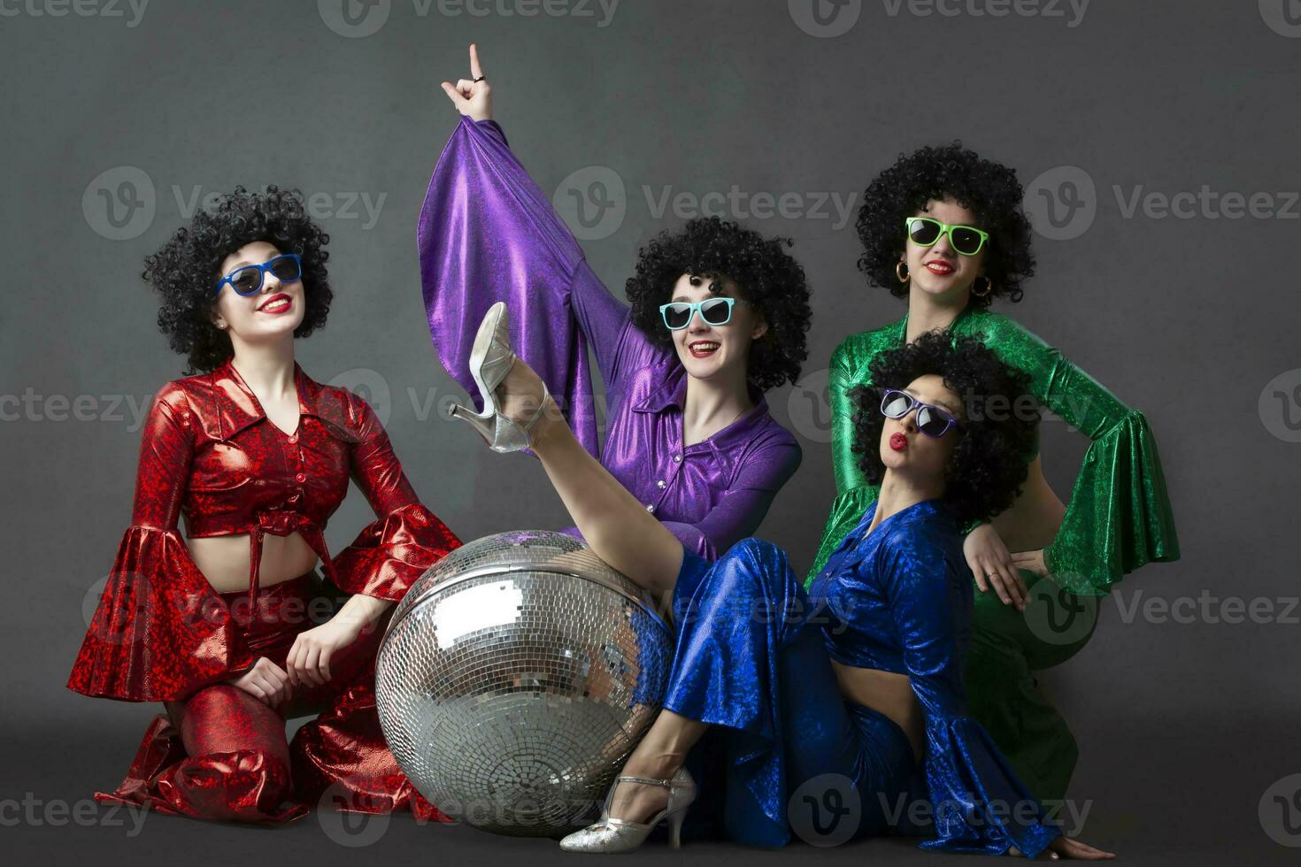 A group of disco girls in wigs with a disco ball and colorful costumes pose against a gray background. Disco party. photo