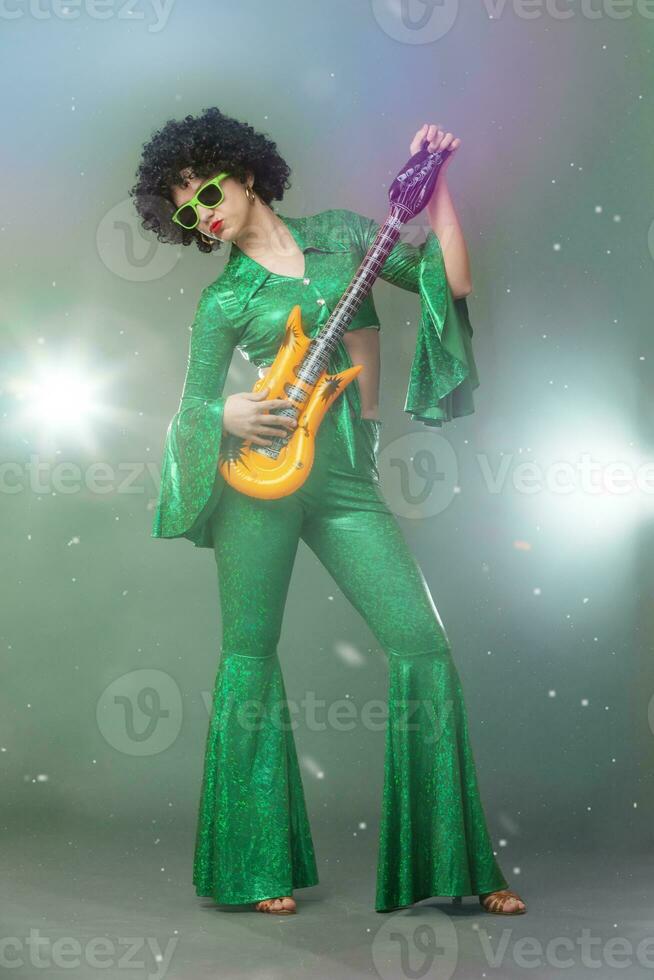 A full-length girl in a flared disco-style stage costume holds a comic guitar in her hands. photo