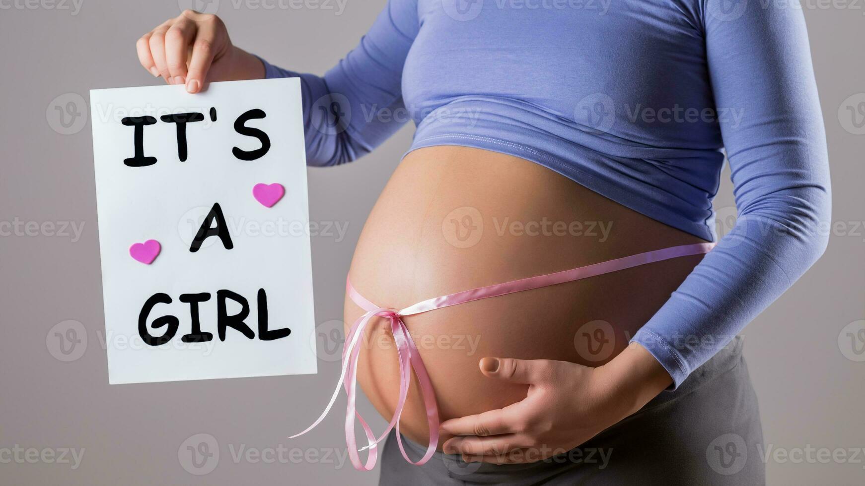 Image of close up stomach of pregnant woman with pink ribbon holding paper with text it's a girl on gray background. photo