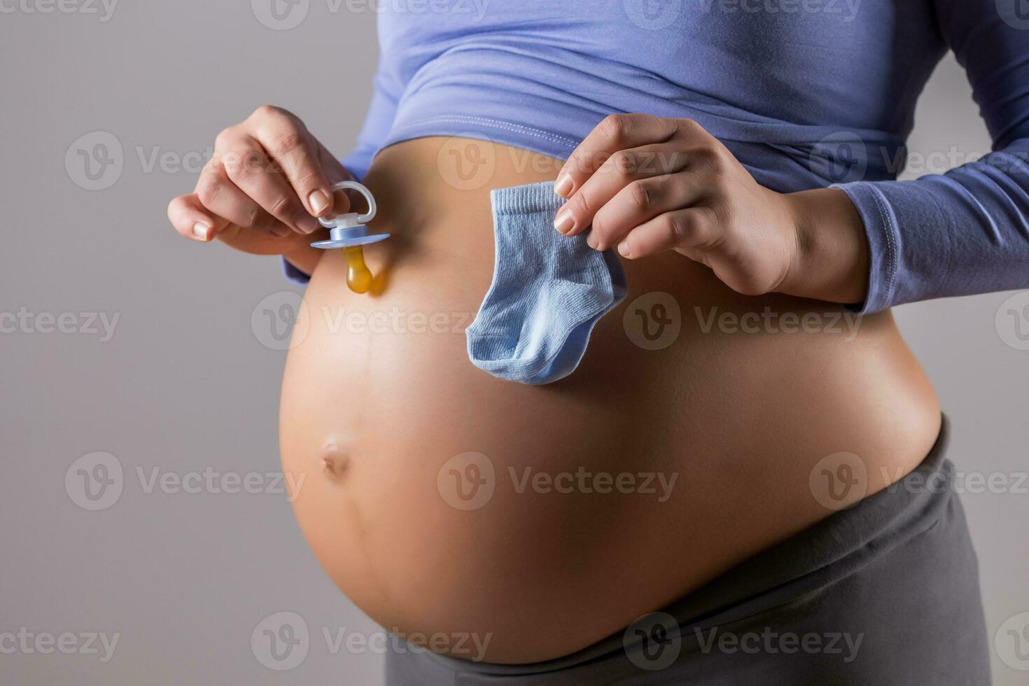Image of stomach of pregnant woman with holding pacifier and socks for baby boy on gray background. photo