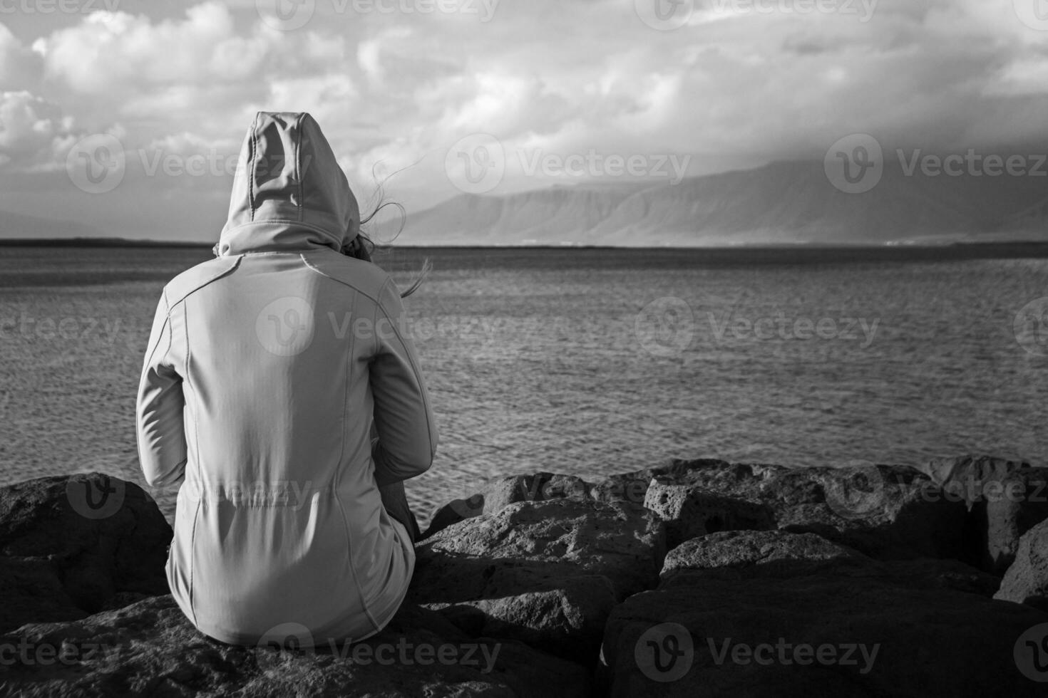 Black and white image of woman sitting at the coastline in Reykjavik and looking at the sea.Image contains little noise because of high ISO. photo