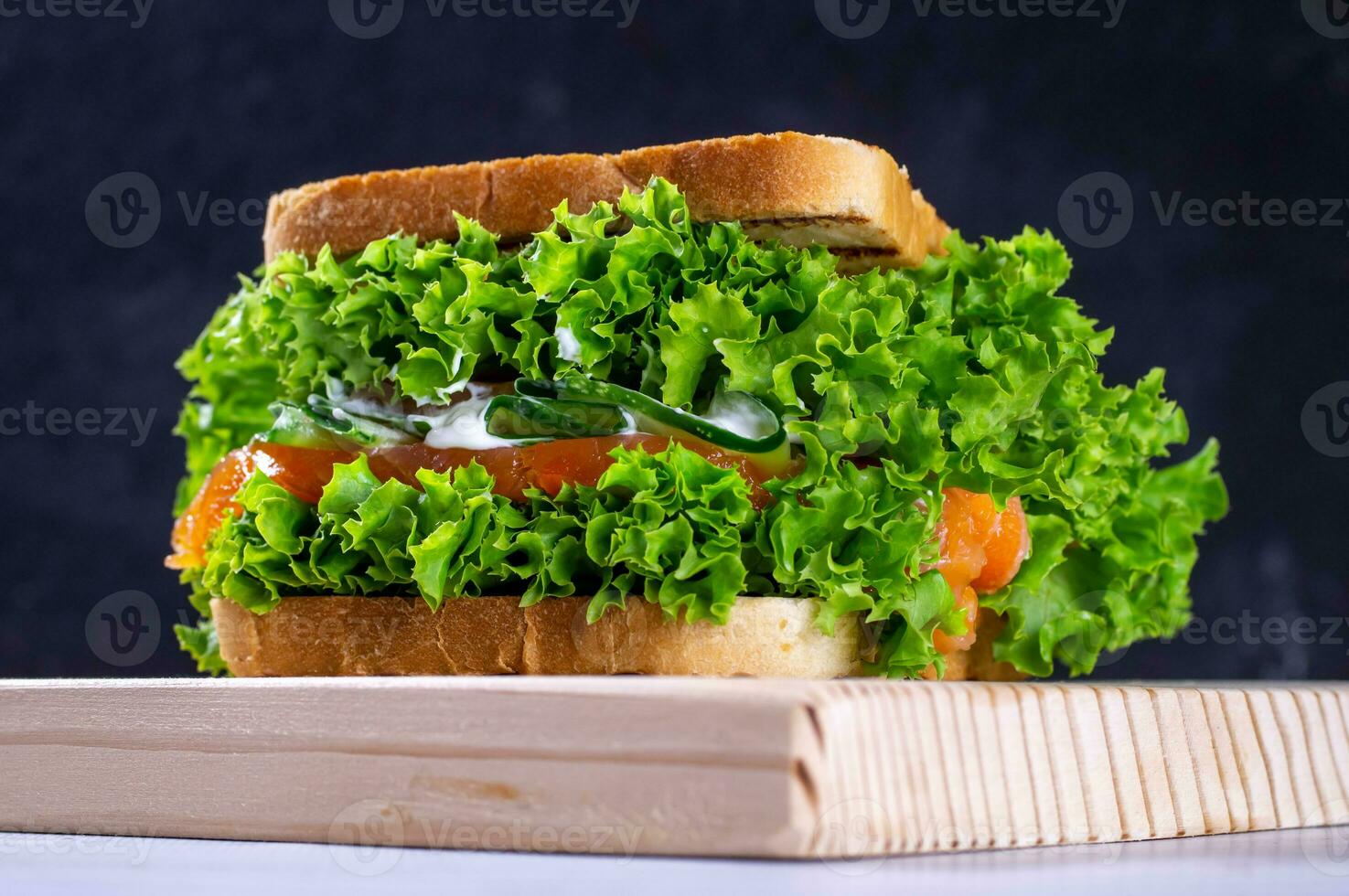 Sandwich with fresh salmon, lettuce, cucumber and sauce on a cutting board, on a dark background. Homemade. Healthy convenience food. photo