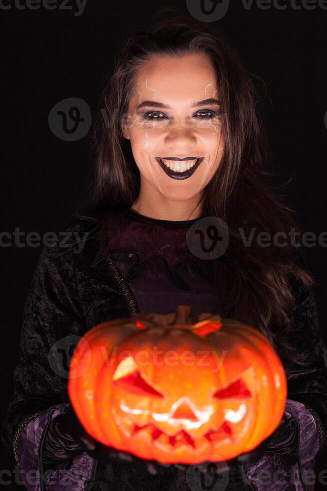 Beautiful woman wearing witch outfit for halloween holding a spooky pumpkin over black background. photo