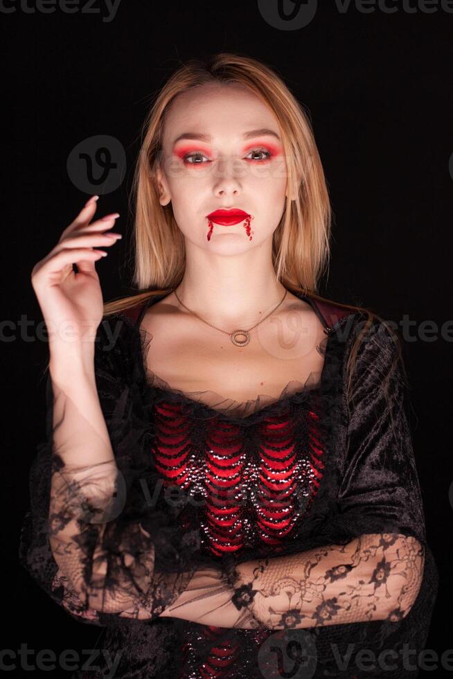 Blond woman dressed up like a vampire with blood on his lips over black background. Halloween costume. photo