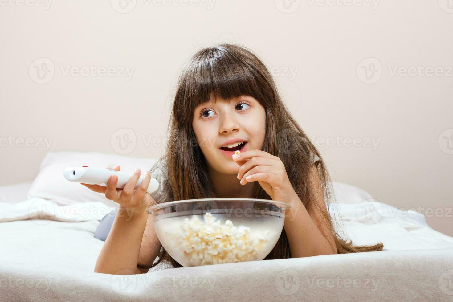 pequeño niña es acecho televisión y comiendo palomitas de maiz en su cama. foto