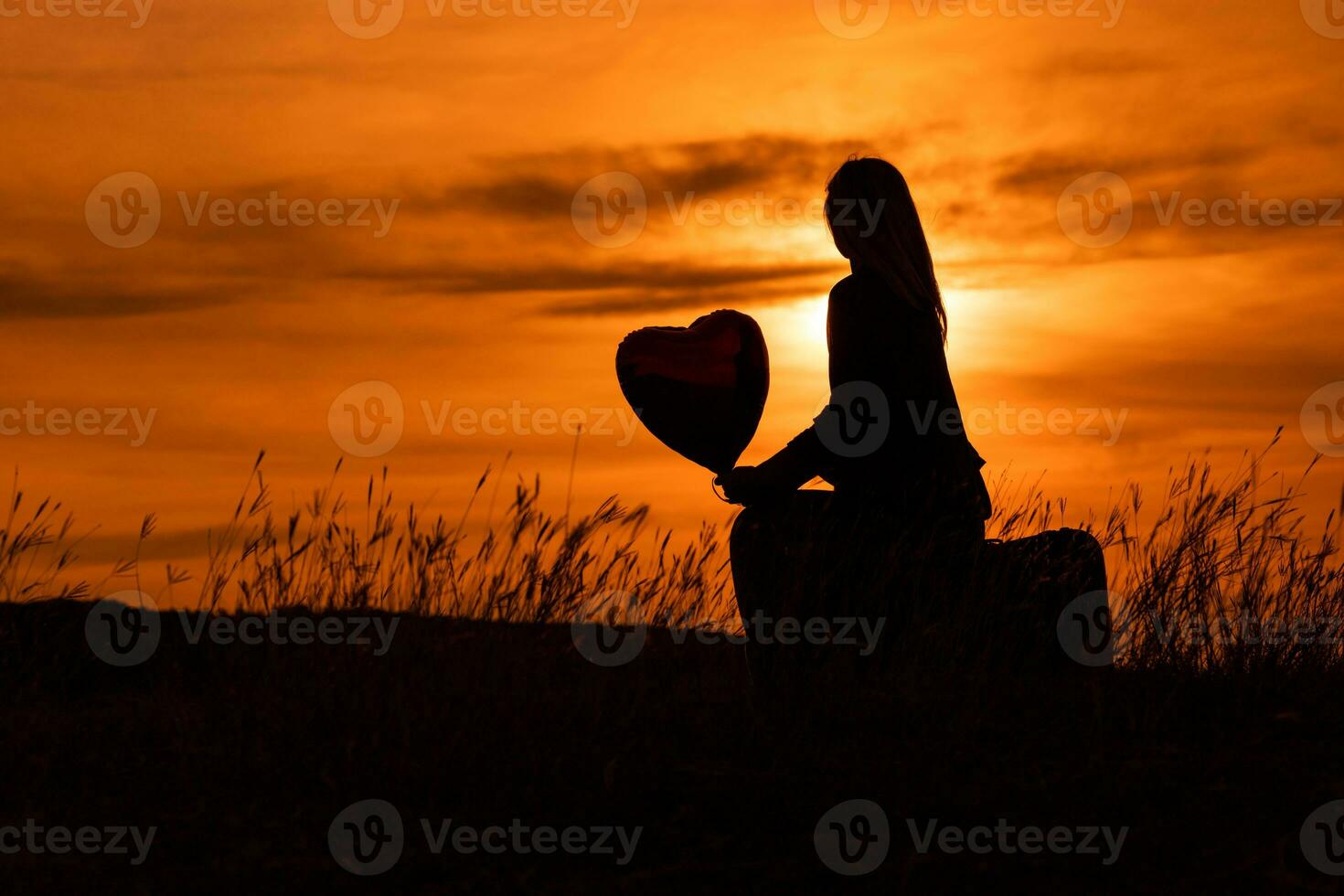 silueta de un mujer sentado en maleta con corazón conformado globo y mirando a hermosa puesta de sol. foto