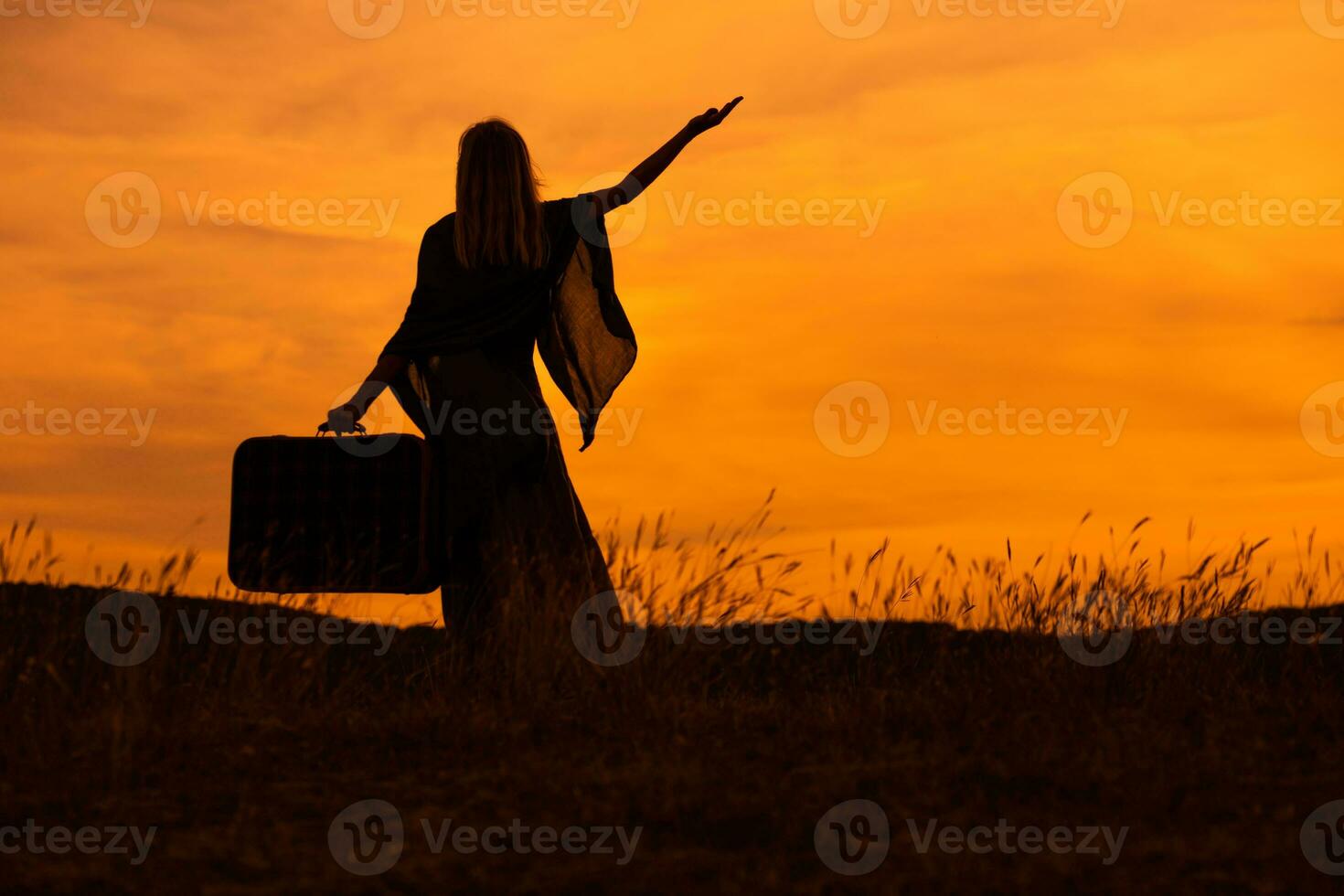 Silhouette of a woman holding suitcase with arm raised and looking at beautiful sunset. photo