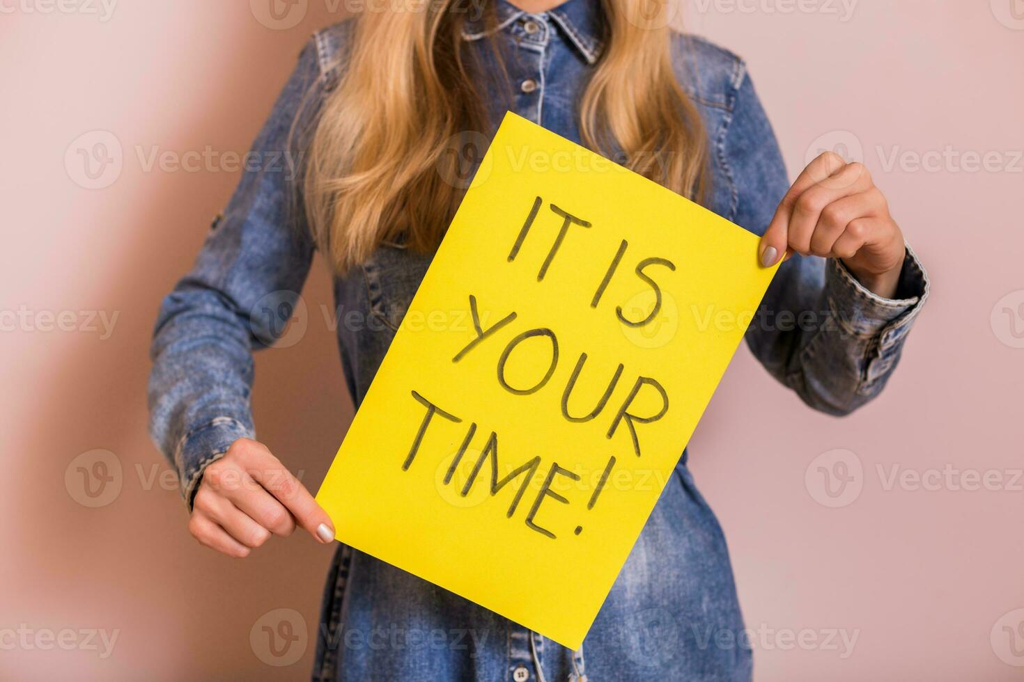 Woman holding paper with text it is your time while standing in front of the wall. photo