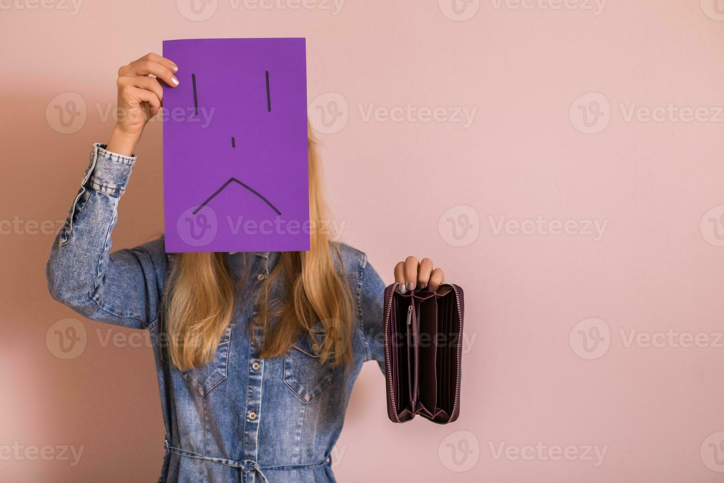 Woman holding empty wallet and sad face on paper while standing in front of wall. photo