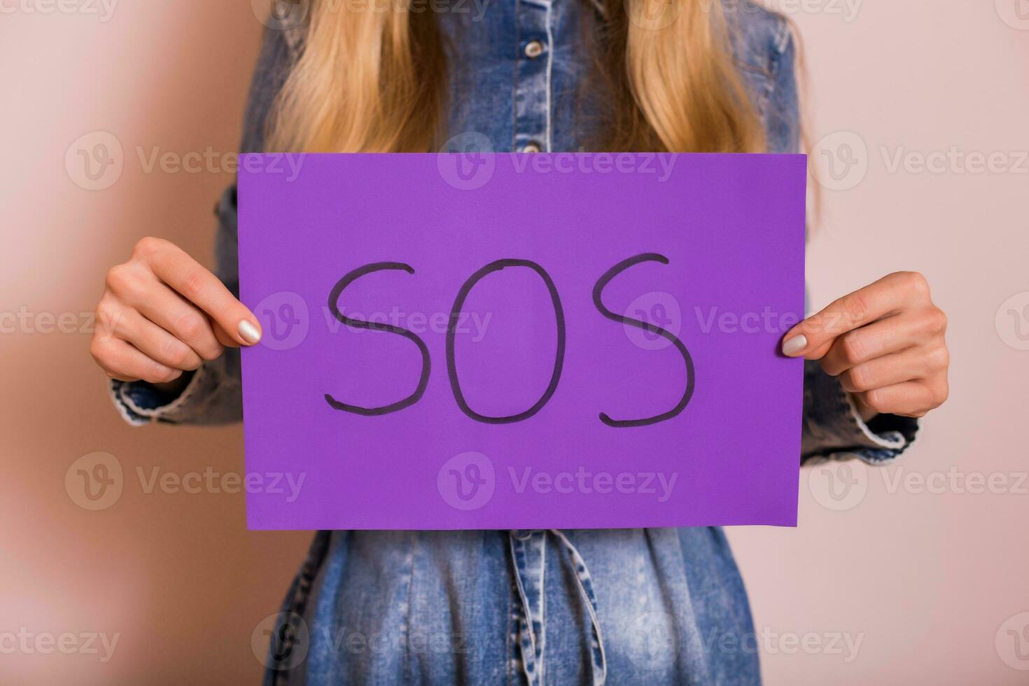 Woman holding paper with word SOS while standing in front of the wall. photo