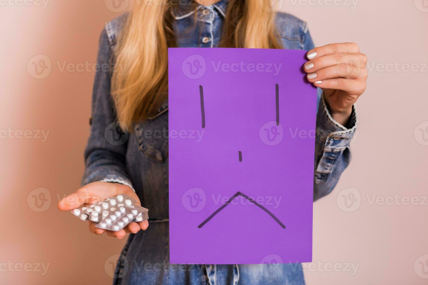 Woman holding pills and sad face on paper while standing in front of wall. photo
