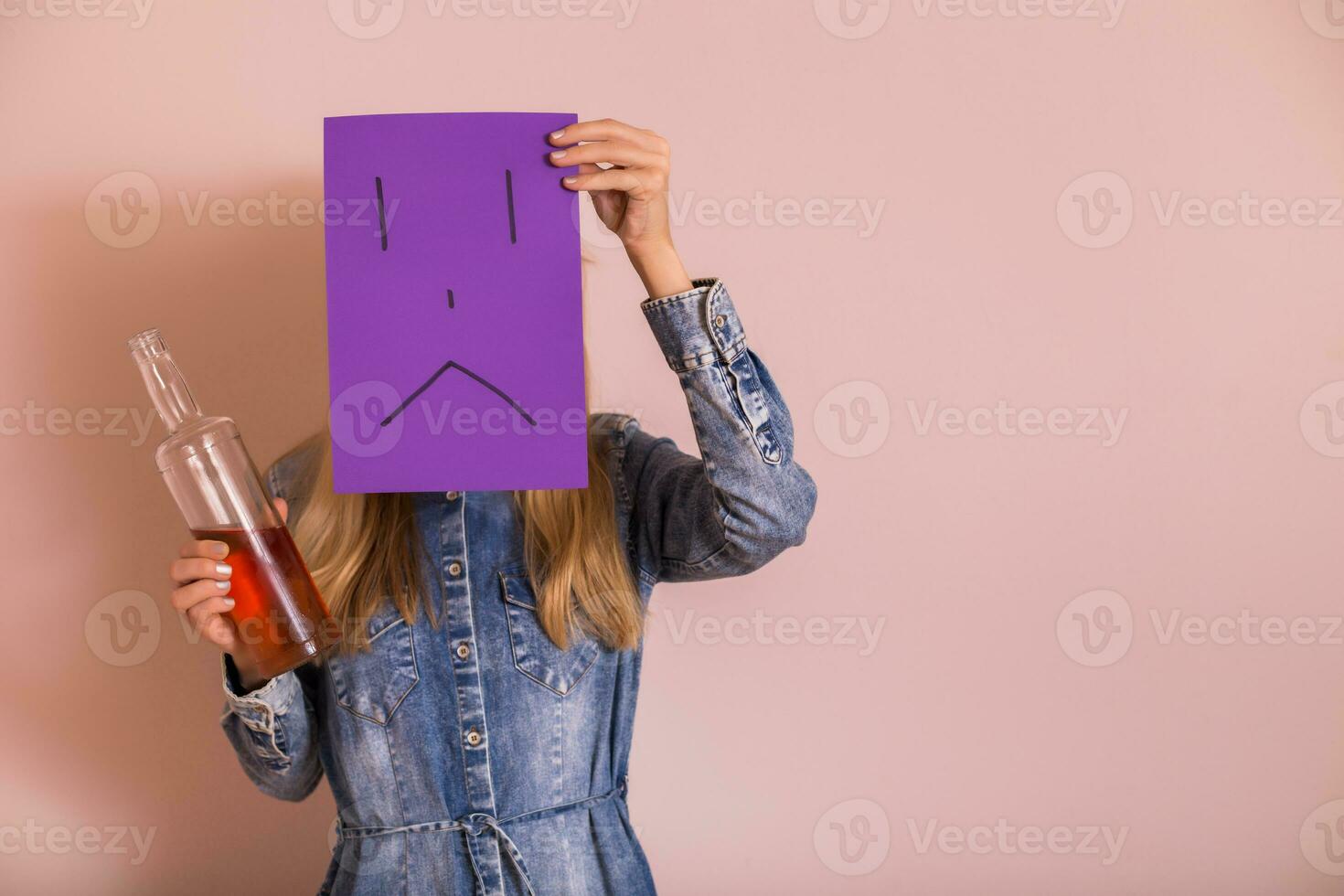 Woman holding alcohol and sad face on paper in front of the wall.Quitting drinking alcohol concept. photo