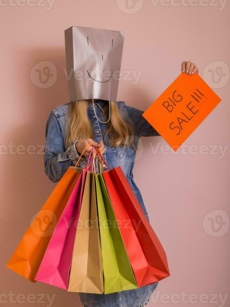 juguetón mujer participación compras pantalones con bolso en su cabeza y papel con texto grande rebaja mientras en pie en frente de el pared. foto