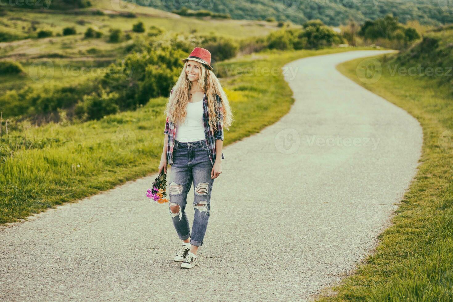 hermosa joven mujer participación ramo de flores de flores y caminando a el país la carretera. foto
