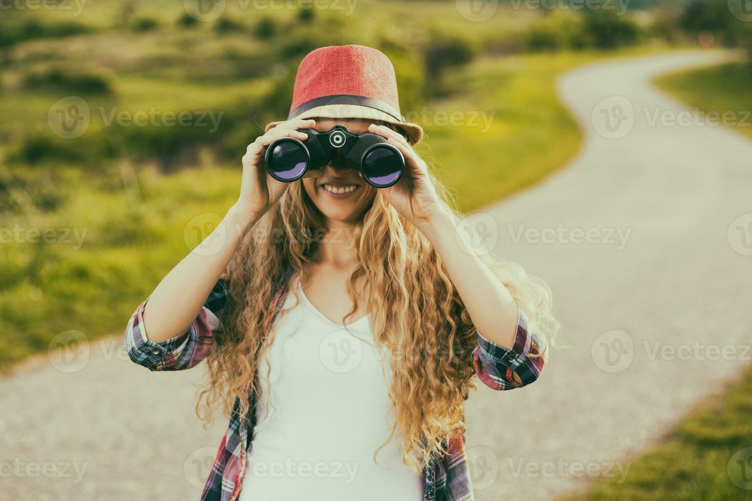 Beautiful young woman at the country road enjoys looking through binoculars.Toned image. photo