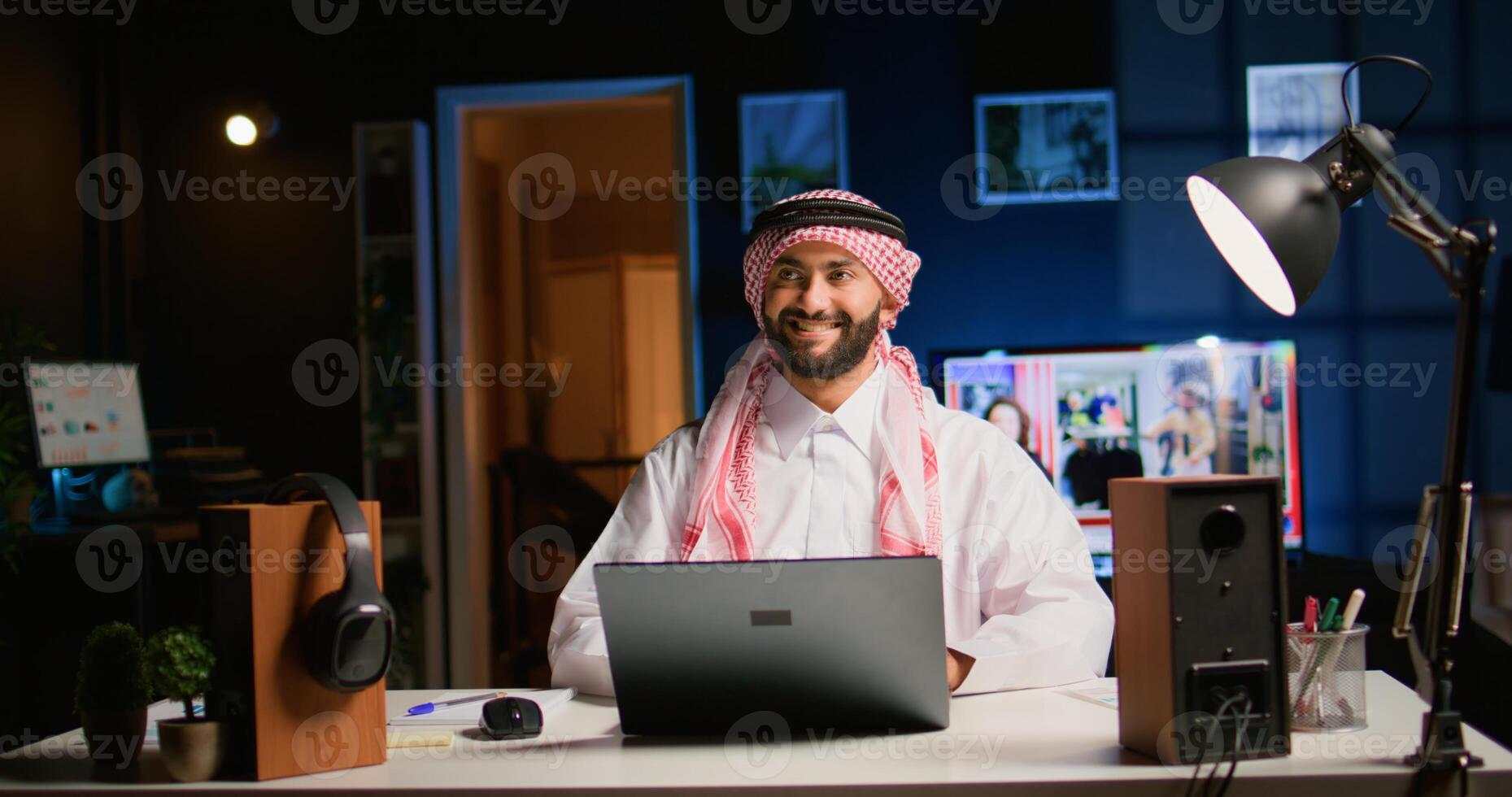 Smiling arab teleworker answering job emails in stylish apartment, happy about finishing work soon. Cheerful muslim employee remotely working, typing data on laptop with opened tv as background noise photo