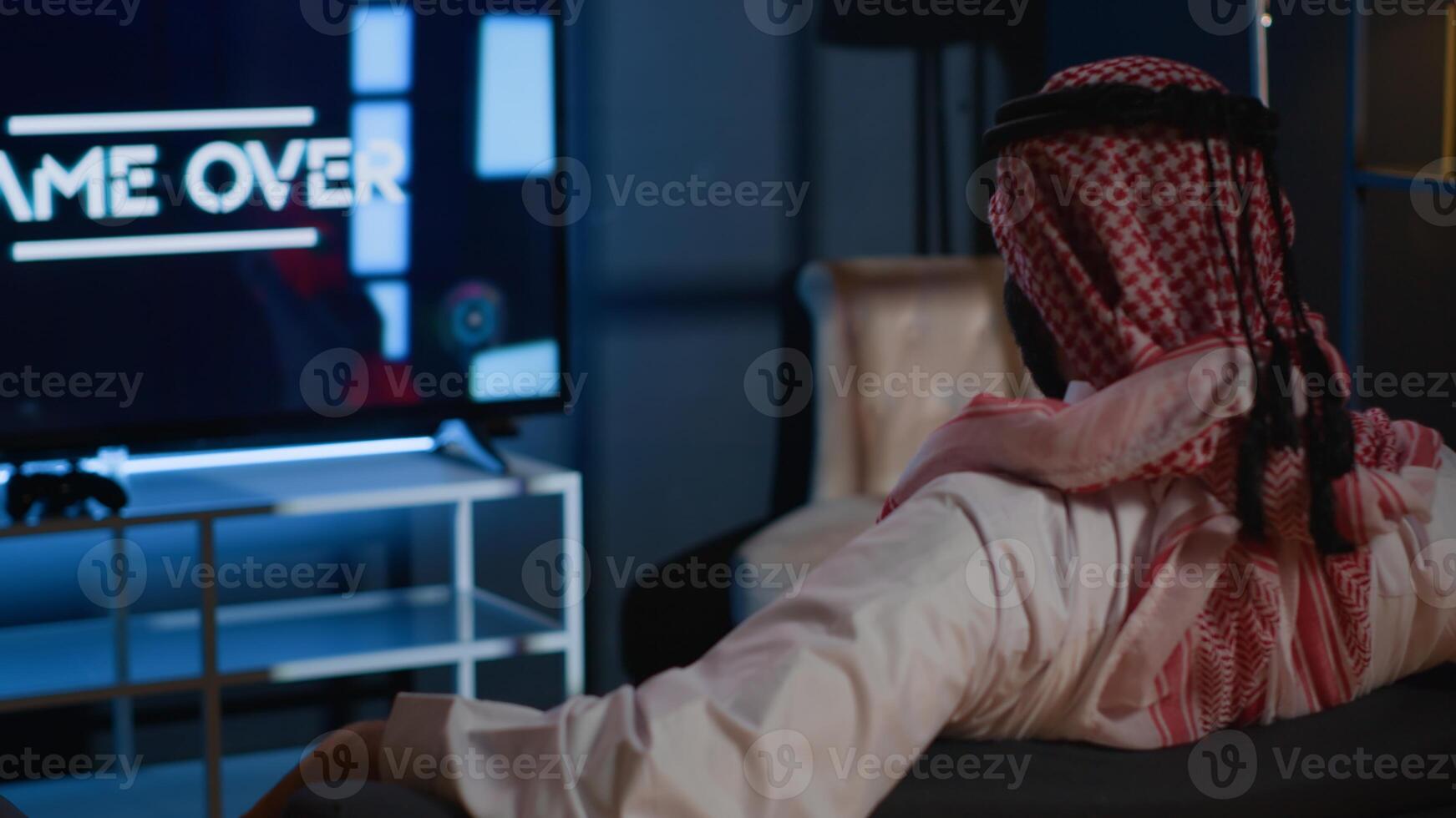 Middle Eastern man playing videogames on TV, relaxing after long day at work. Competitive gamer upset after losing science fiction shooter game on gaming console in stylish apartment living room photo