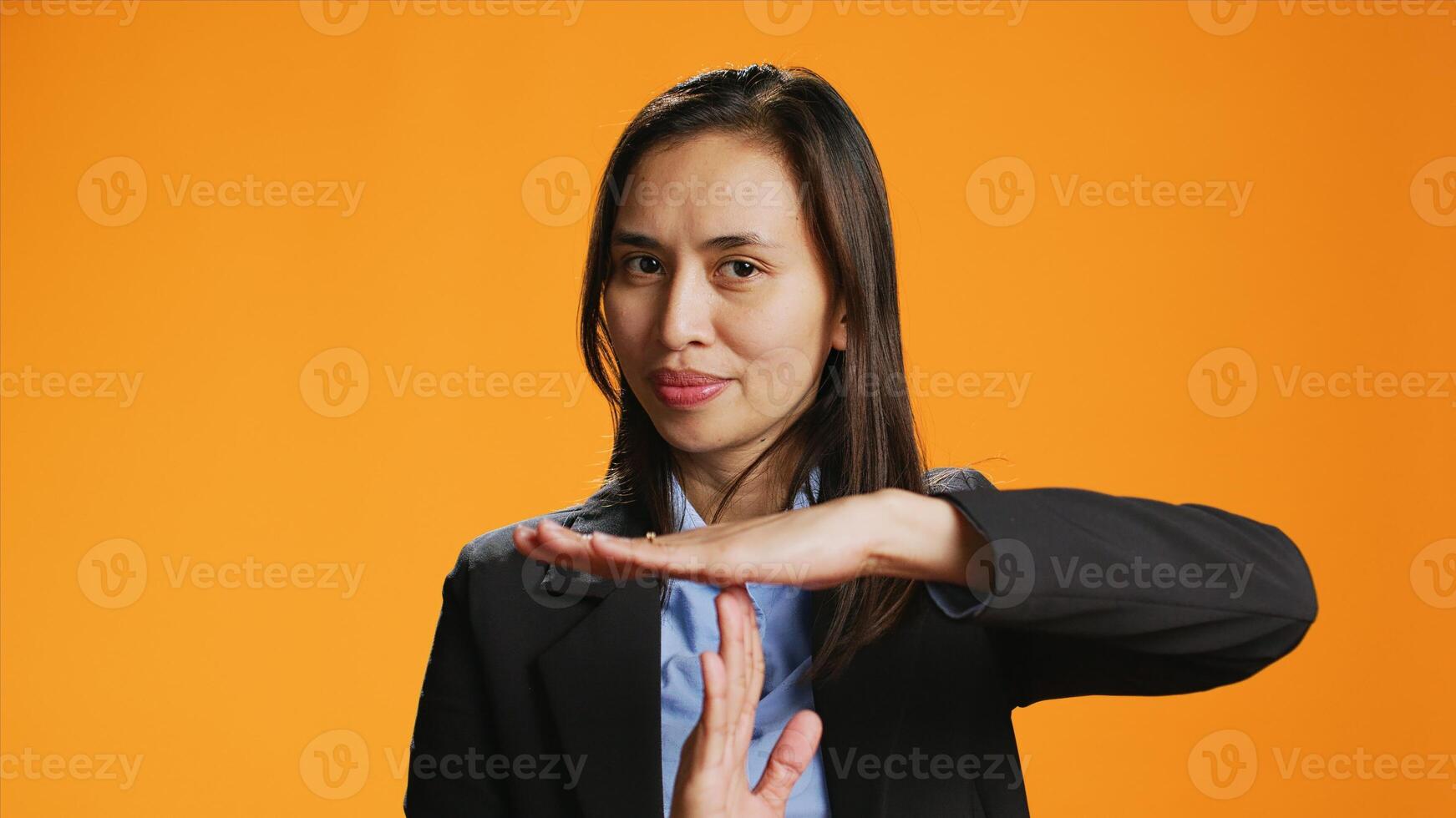 Serious adult presenting timeout gesture in studio, asking for a break after working too hard at the office. Asian woman standing over orange background, doing pause symbol with hands. photo