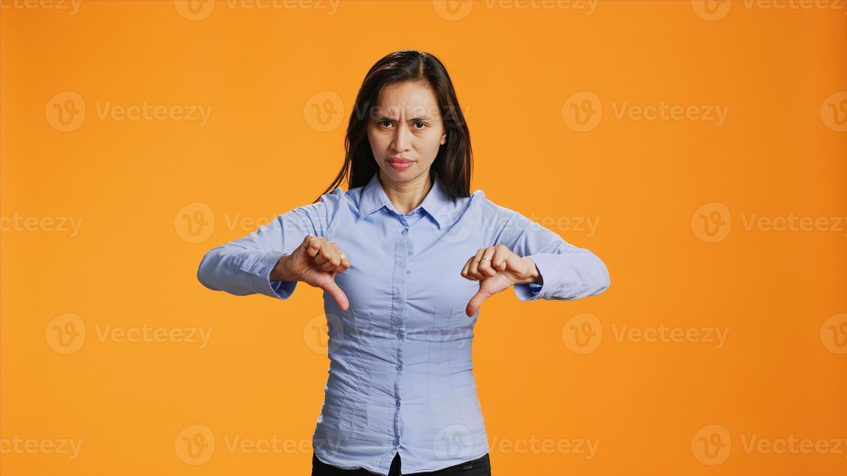 Asian woman giving dislike gesture on camera, showing disagreement and negativity over orange background. Negative unhappy adult presenting thumbs down symbol in studio, wrong idea. photo