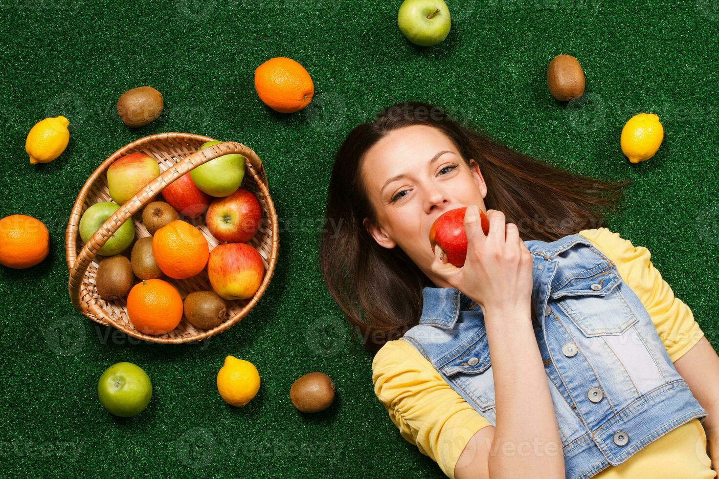 Beautiful young woman is eating apple while lying down on the grass with a lot of fruit around her photo