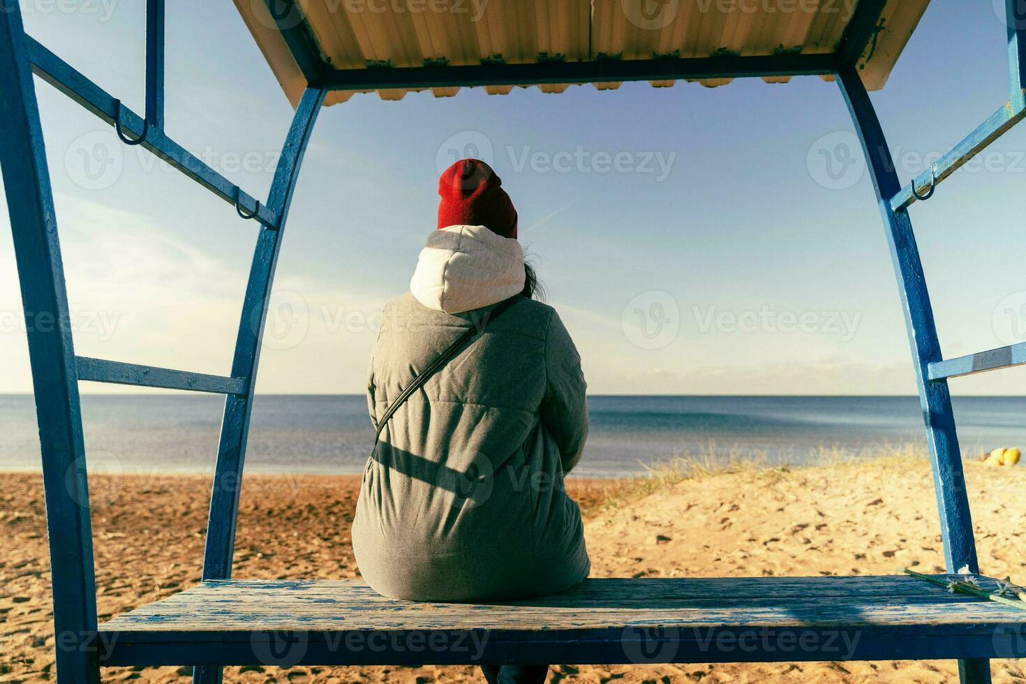 woman in an autumn jacket and hat is sitting alone on a bench on a deserted autumn beach on a sunny day photo