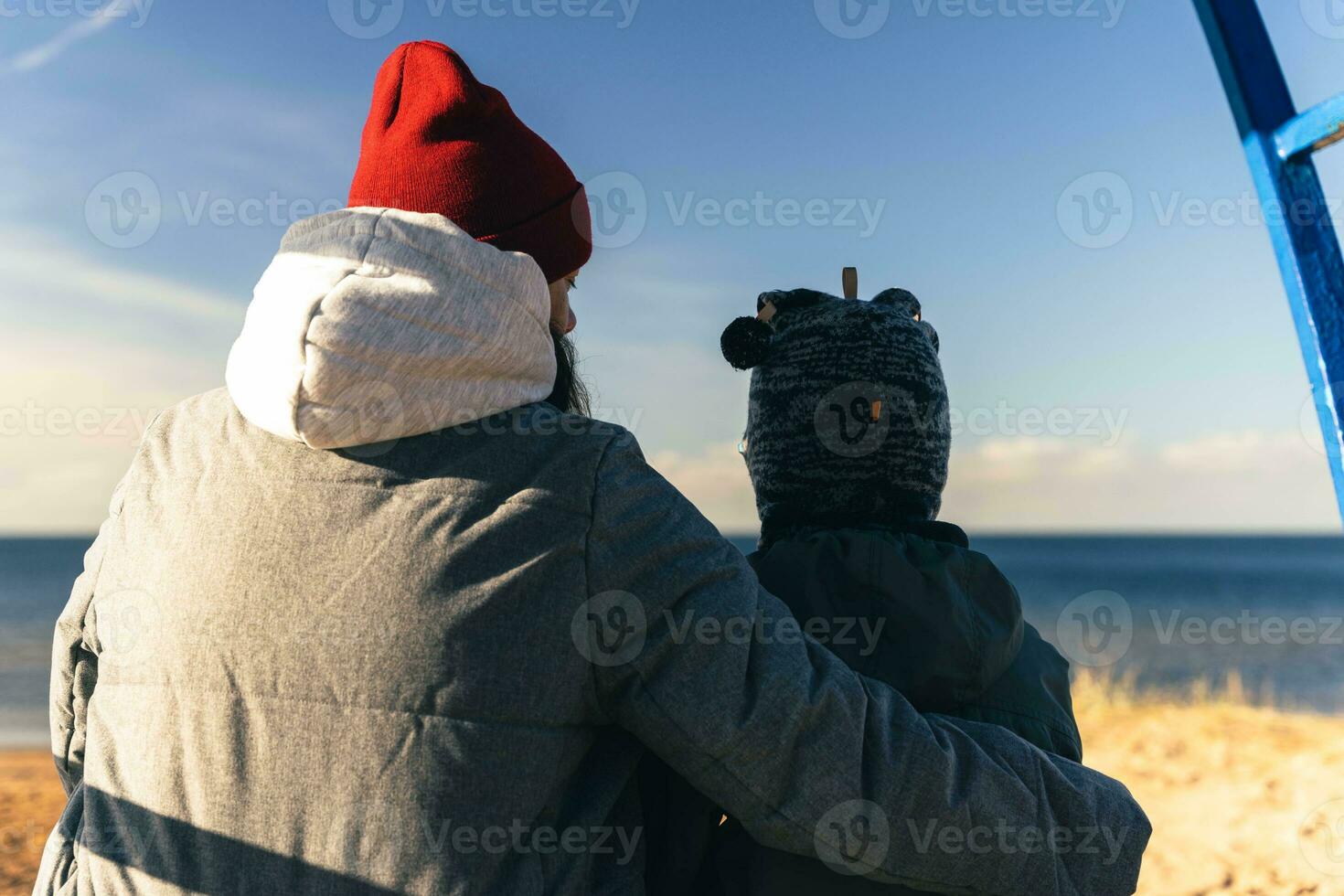 woman in an autumn jacket and her child are sitting on a bench on a deserted autumn beach on a sunny day photo