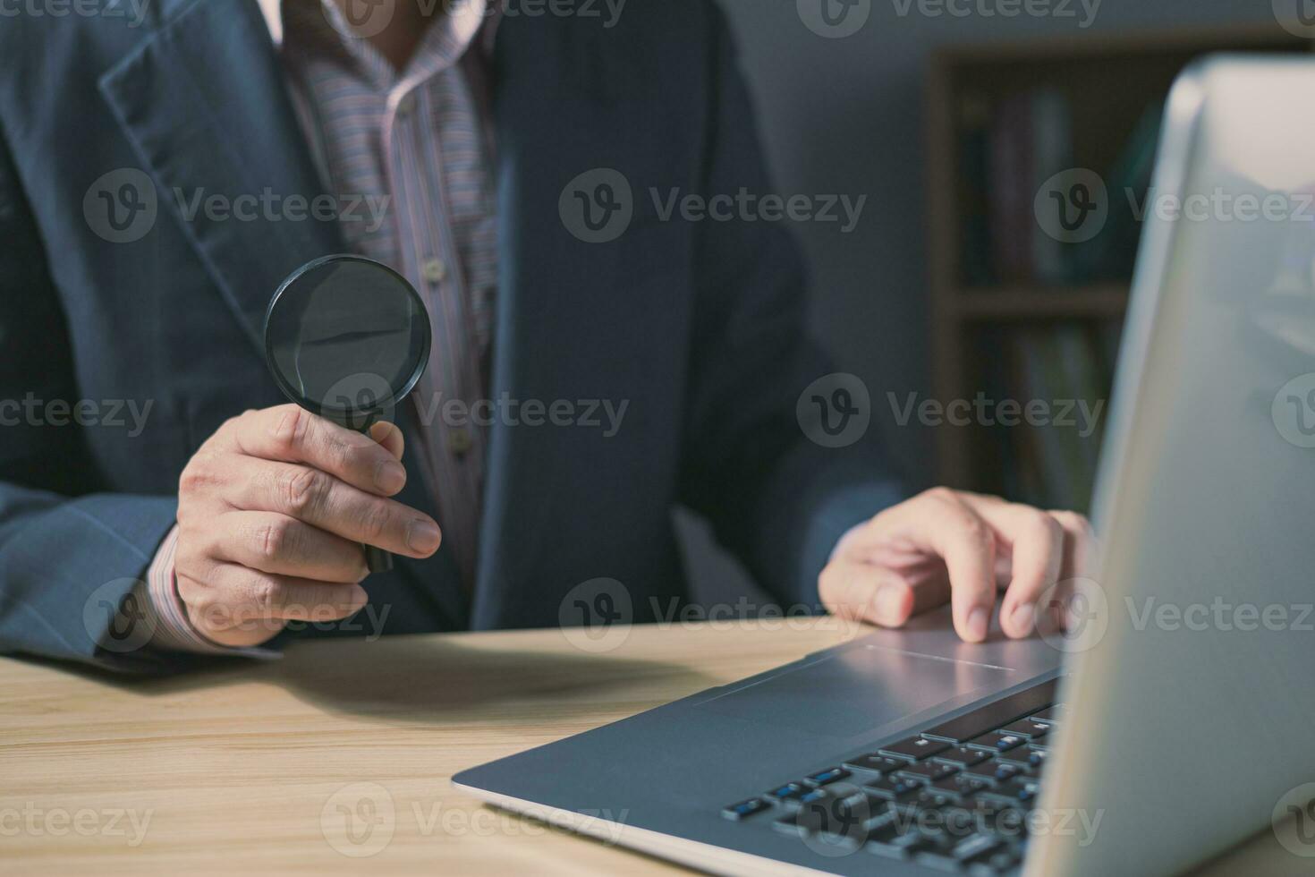 Businessman sit at office in suit holding magnifying glass and computer laptop.Search and research concept. photo