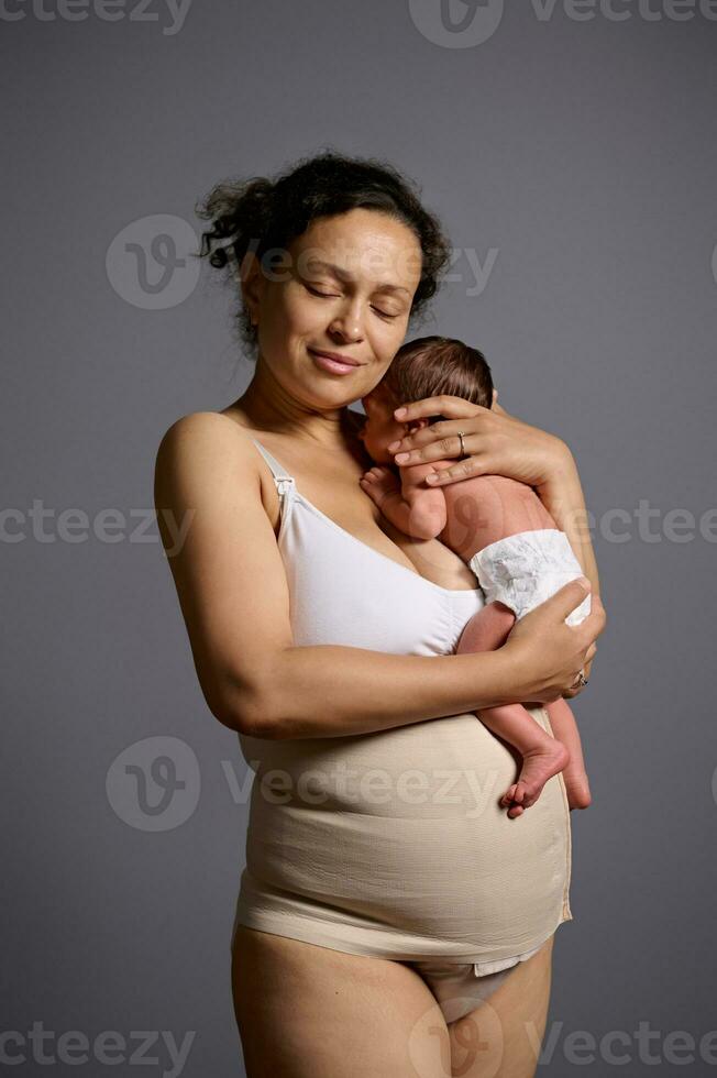 amoroso cuidando multiétnico joven mujer madre que lleva y abrazando su recién nacido bebé, aislado terminado gris estudio antecedentes foto