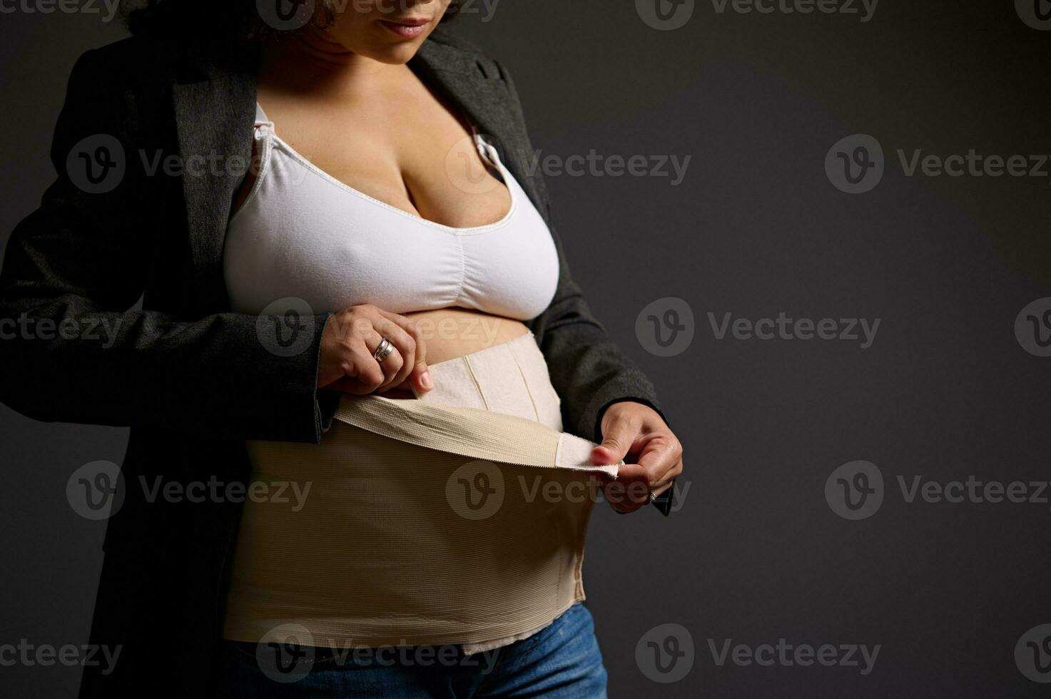 Close-up young woman wearing an elastic bandage on her postnatal belly, isolated on gray background. photo