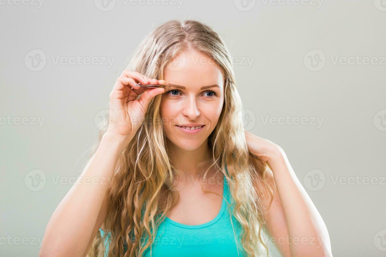 Beautiful young woman plucking eyebrows on gray background. photo