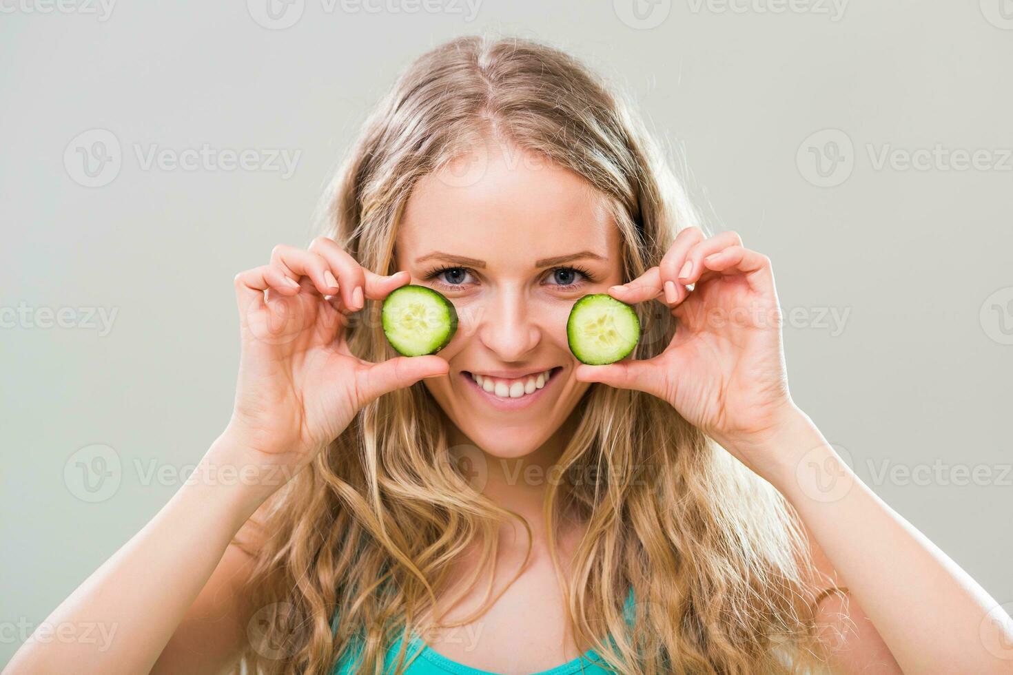 Beautiful young woman showing slices of cucumber on gray background. photo