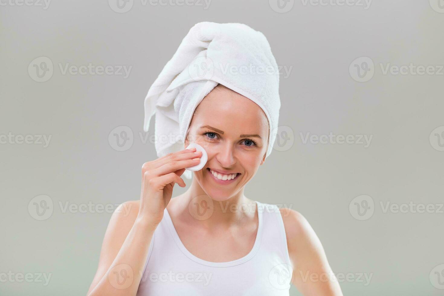 Beautiful young woman cleaning her face with cotton pad on gray background. photo