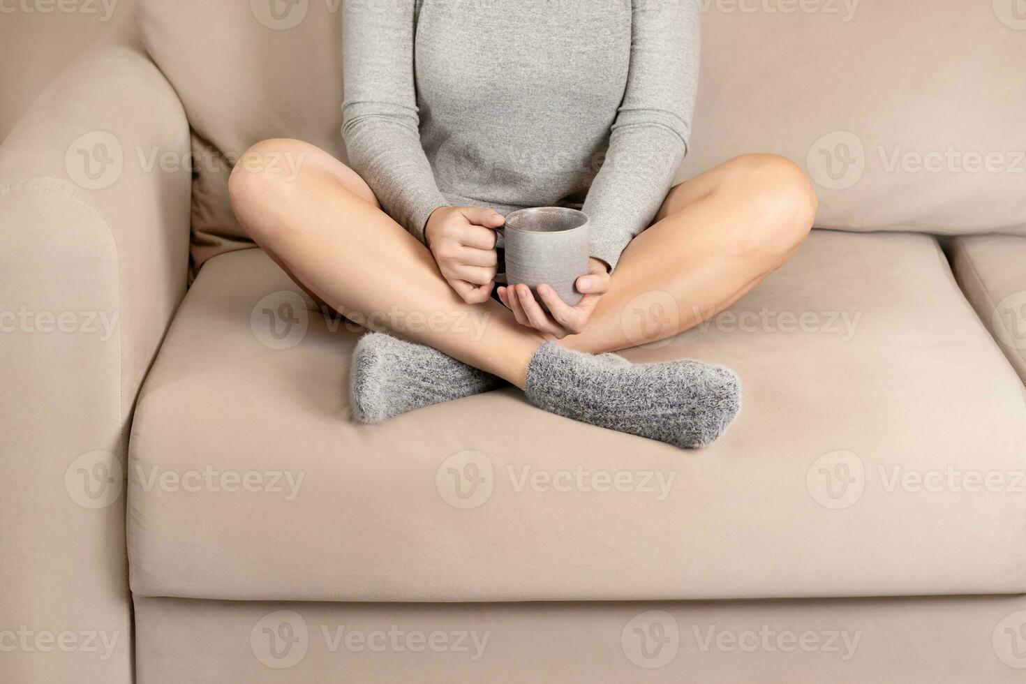 Woman sitting in lotus position on the sofa and drinking hot tea beige grey colors close up cozy photo