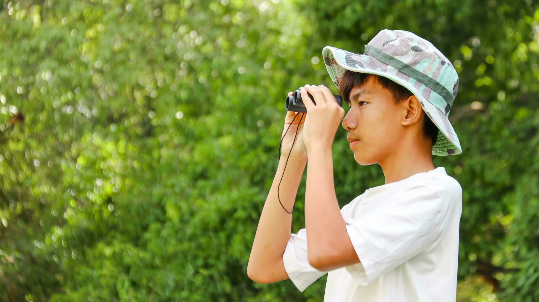 Young Asian boy is using a binocular to lookout for birds and animals in a local park, soft and selective focus photo