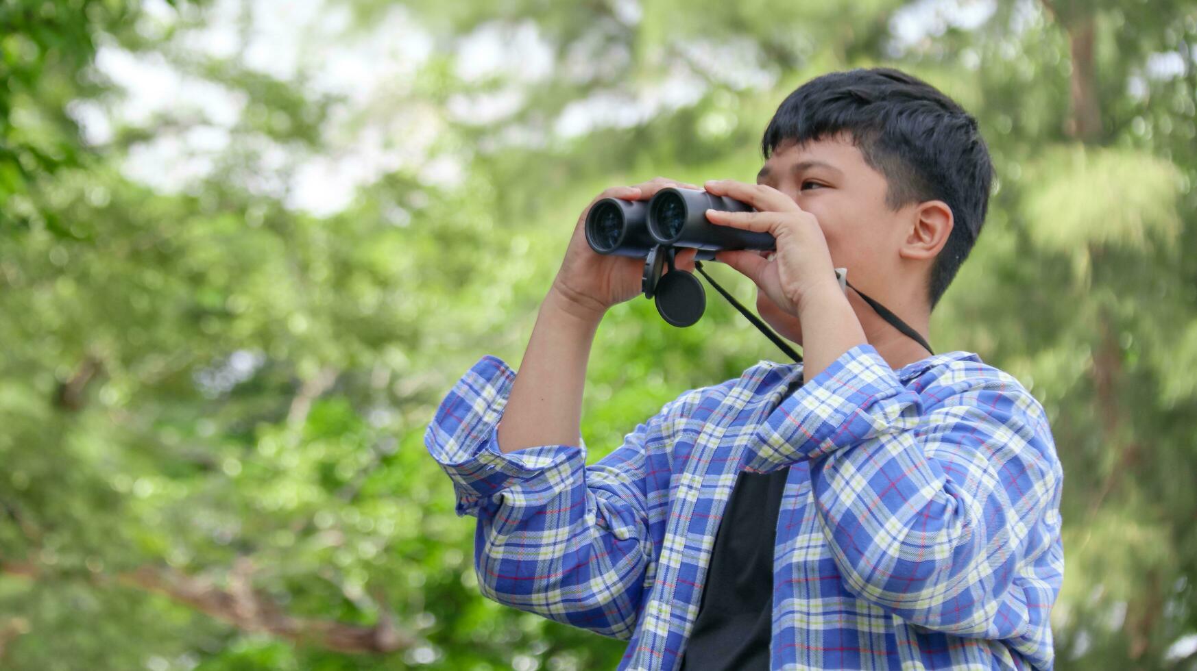 Young Asian boy is using a binocular to lookout for birds and animals in a local park, soft and selective focus photo