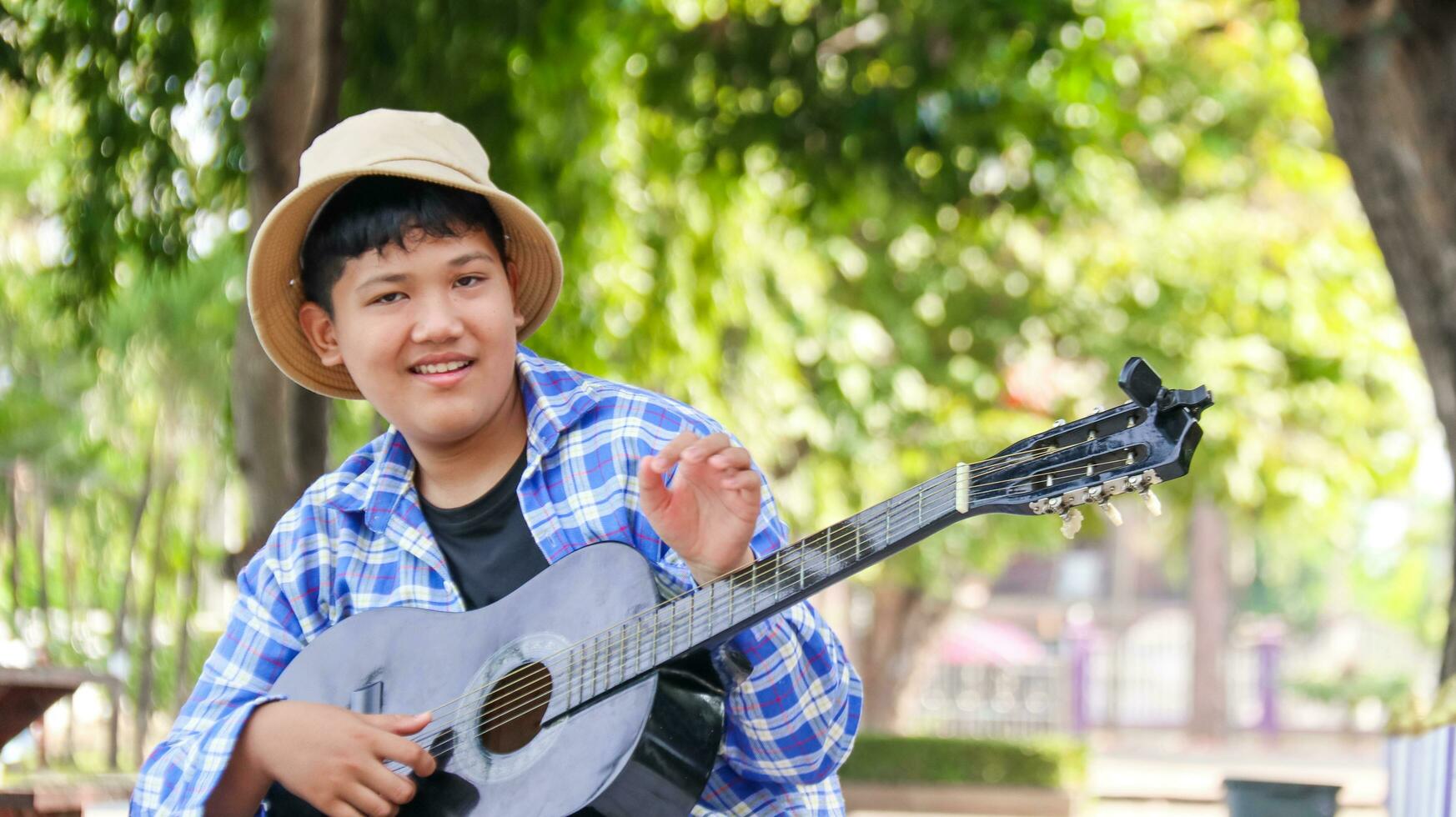 joven asiático chico es jugando guitarra en un local parque, suave y selectivo atención foto