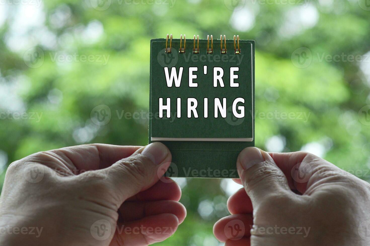 Hands Holding notepad with we are hiring text against blurred green background. photo