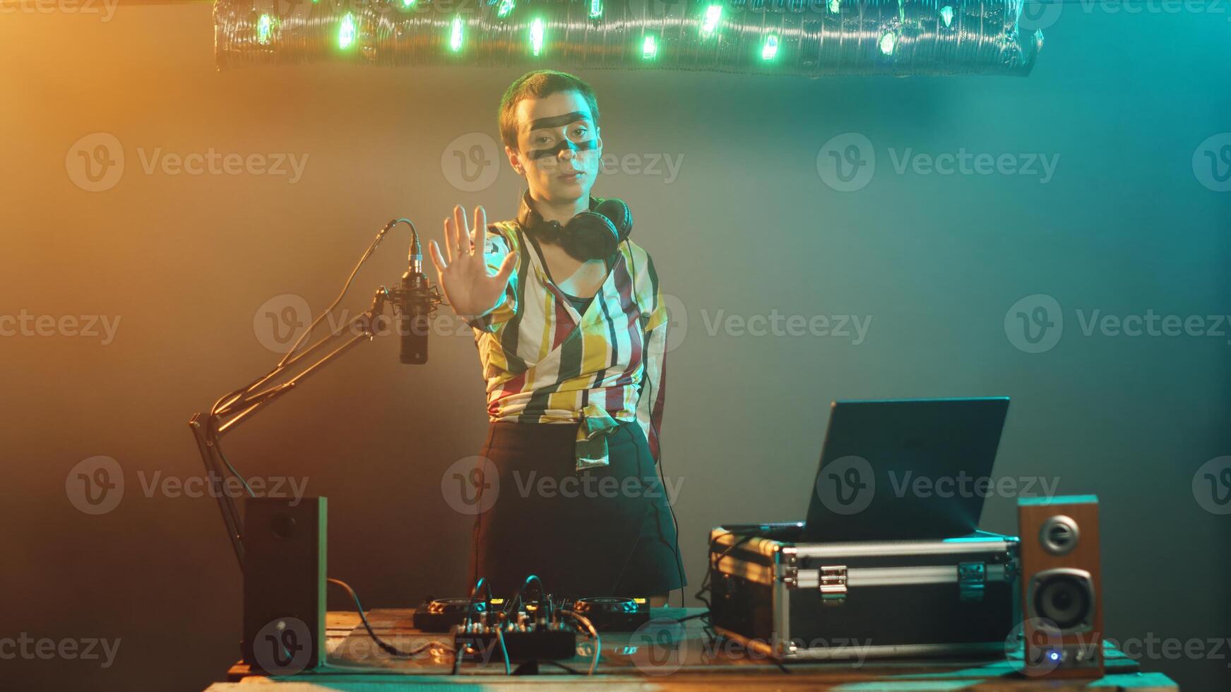 Crazy club musician raising palm to show denial and stop symbol on stage, mixing modern techno sounds on DJ turntables. Performer using audio stereo instruments, adjusting buttons. Tripod shot. photo