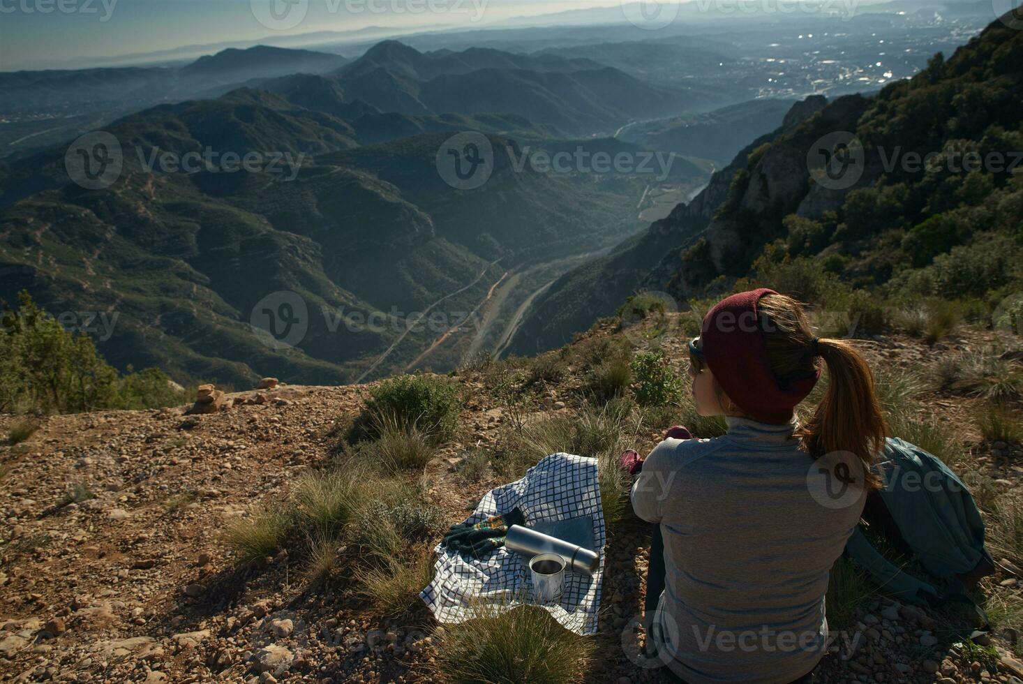Woman takes a break at the top of the mountain observing the landscape, with a thermos and a cup of coffee. photo