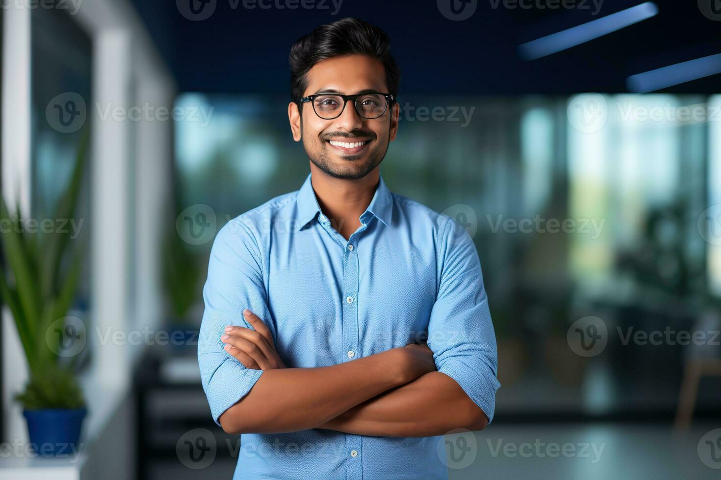 ai generado un joven indio masculino diseñador, ingeniero, arquitecto quien es vistiendo lentes y un azul camisa sonriente en pie en el oficina y mirando a el cámara foto