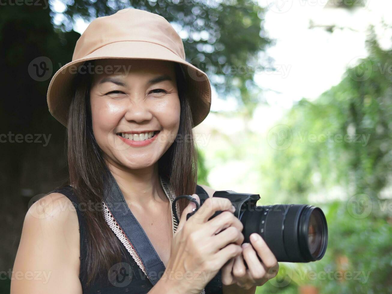 Asian woman, wearing hat and black top sleeveless, standing in the garden and  holding dslr camera, smiling happily. photo