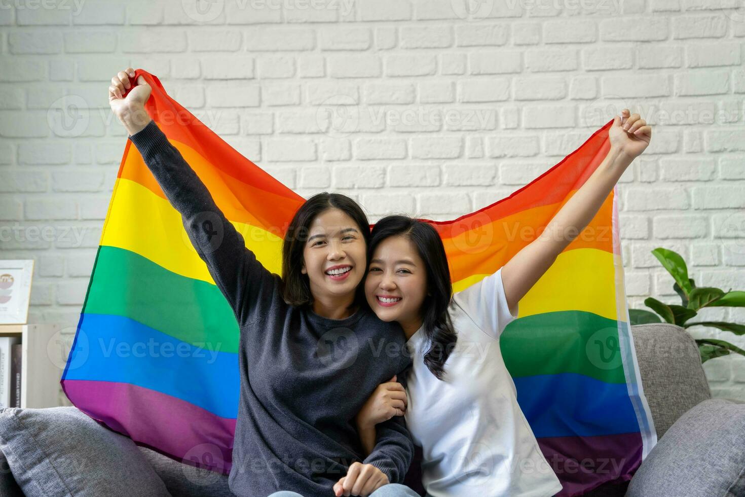 lgbtq orgullo mes, retrato de contento asiático lesbiana Pareja con arco iris banderas sentado en sofá a hogar foto