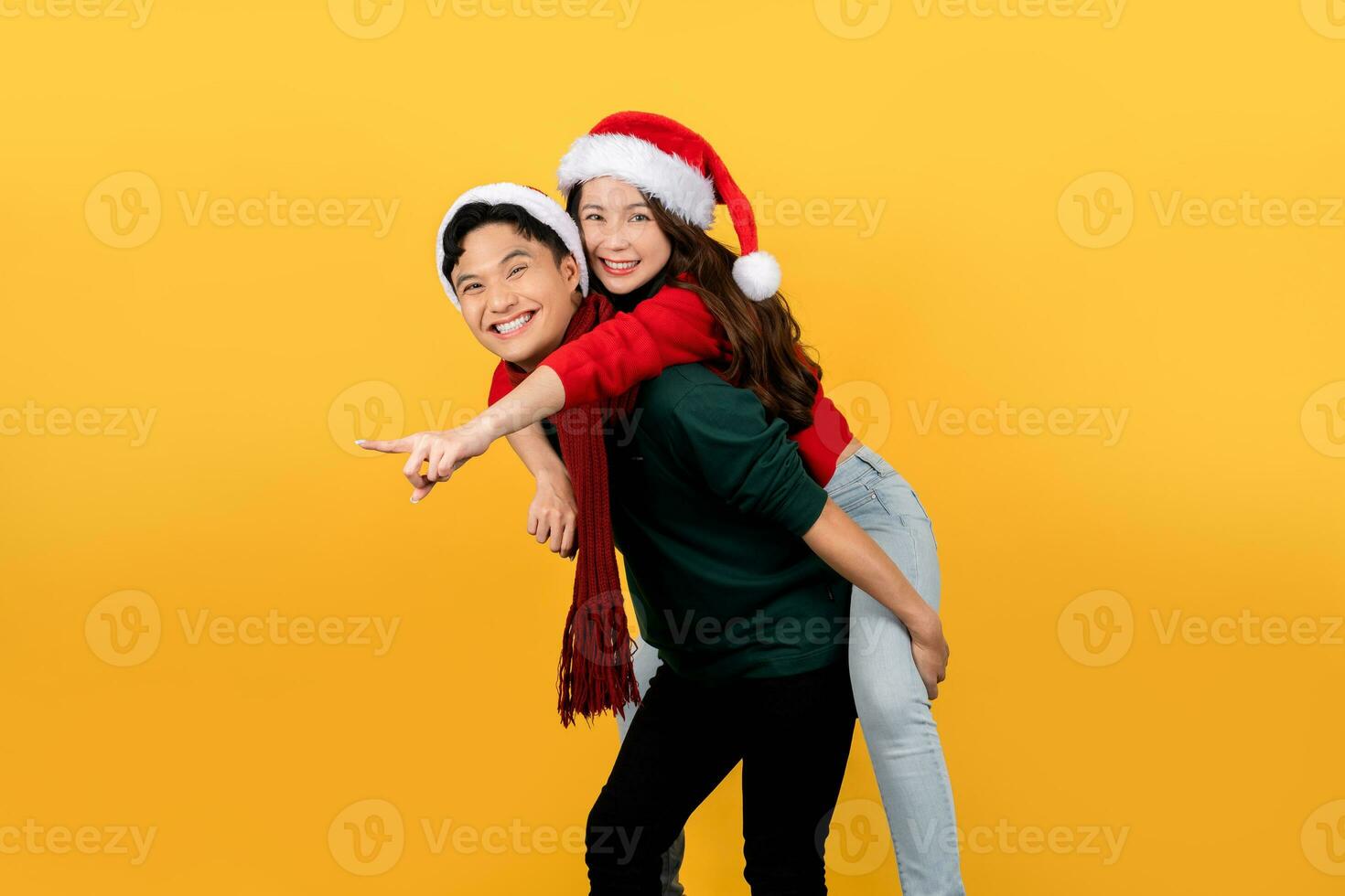 Smiling Asian couple wearing sweaters and Santa hats playfully posing piggyback on a yellow background. Happy New Year or Christmas holiday concept. photo