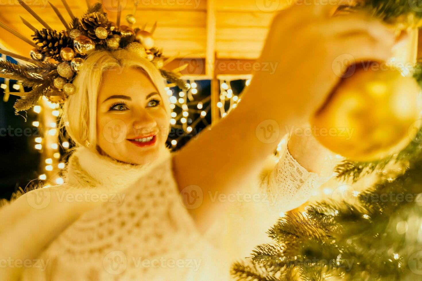 un rubia mujer en blanco vestir y un corona de oro adornos Decorar Navidad árbol con oro adornos y luces. el árbol es decorado con oro pelotas y es iluminado arriba con luces. foto